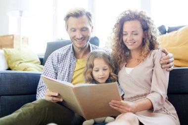 Genç ve sevecen bir çift ve küçük kızları birlikte oturma odasında kitap okuyorlar.