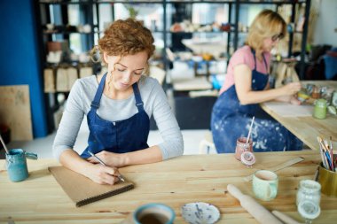 Çalışma kıyafetlerindeki genç yaratıcı çömlekçi masaya oturup notlar alıyor ya da not defterine çizim yapıyor.