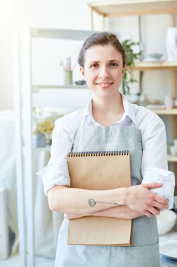 Sanat stüdyosunda kameraya bakan not defteri ile gülümseyen yaratıcı kadın