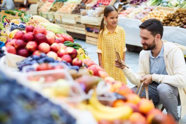 İçerik yakışıklı genç sakallı baba gıda tezgahı yakınında çömelmiş ve kızı ile birlikte organik gıda satın alırken çiftçi pazarında alışveriş listesini kontrol