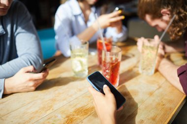 Genç insanlar güneşli bir günde ahşap masada oturup içki içerken akıllı telefonlarını kullanıyorlar.