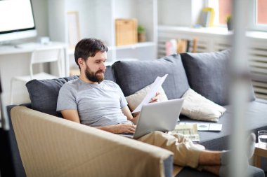 Evde kanepede oturan casualwear rahat adam, kağıtları okuma ve ağ