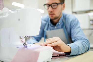 Dikiş makinesinde dikiş makinesinde masada oturan ve dikiş kumaşı diken konsantre genç adam, terzilik stüdyosunda çalışıyor