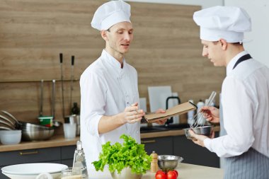 Şapka tutan not defteri ve pişirme sınıfında çırpma ve kase ile stajyer öğretirken tarifi açıklayan emin yetenekli şef