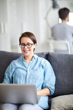 Neşeli genç ev ofis yöneticisi dizüstü önünde kanepede oturan ve web siteleri ile çalışan