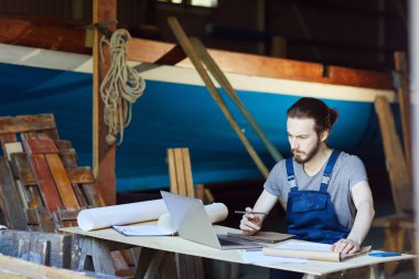 Önlüklü genç mühendis dizüstü bilgisayarın önünde oturuyor ve çizim yaparken görüntüsüne bakıyor.