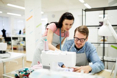 Atölyede dikiş makinesinin nasıl kullanılacağını stajyeranlatırken boynuna santimetre boyunda ciddi yetenekli genç bayan terzilik konusunda rehberlik ederken