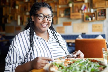 Afrika örgülü mutlu genç kadın masa başında otururken ve öğle yemeğinde pizza yerken size bakıyor.