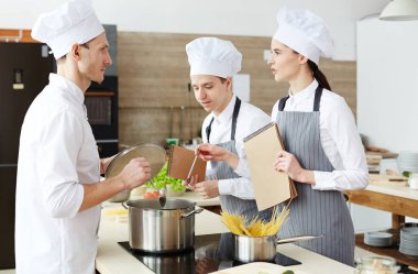 Öğretmen ile makarna tarifi tartışırken pratik sınıfta sorular soran şefler şapka ve önlük meraklı genç pişirme öğrencileri