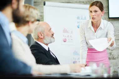 Scowling businesswoman in white shirt and pink skirt looking at one of colleagues while making report at seminar clipart