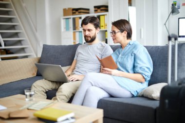 Genç eşler kanepede oturur ve uygun birini seçerken internetteki sıcak tur tekliflerine bakarlar.