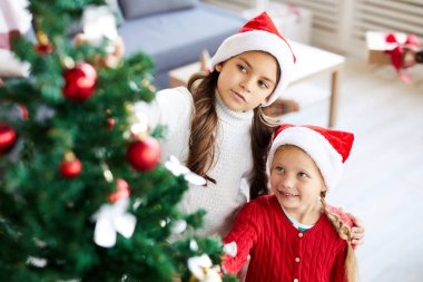 Noel Baba şapkalı iki küçük kız noel ağacının dekorasyonlarına bakarken yanında dururken