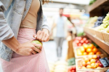Close-up of unrecognizable woman in stripped skirt hiding apple in pocket while stealing it in food store clipart