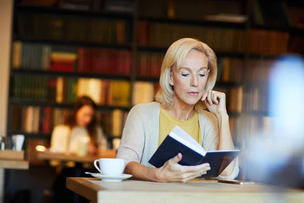 Pensive Blonde Mature Woman Sitting Desk Library Cafe Reading Book — Stock Photo, Image