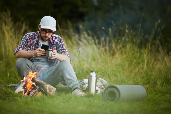 Genç Adam Çay Içiyor Kamp Ateşinin Yanında Akıllı Telefonunu Kullanıyor — Stok fotoğraf