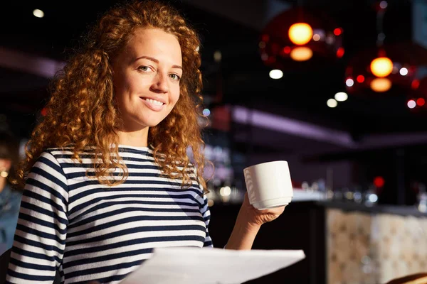 微笑迷人的年轻女子 卷曲的头发拿着文件 喝着咖啡 同时看着相机在现代咖啡馆 — 图库照片