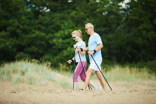 现代高级夫妇戴着太阳镜和活动服徒步在沙滩上跋涉 背景是绿树 — 图库照片