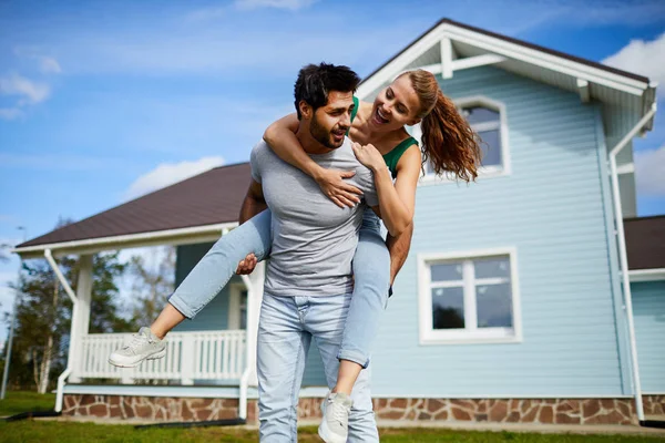 彼らの新しい家の背景に彼の陽気なガールフレンドのピギーバックを与える若い男 — ストック写真