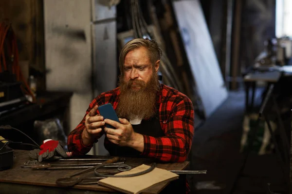 Frowning Puzzled Bearded Blacksmith Checkered Shirt Using Smartphone While Checking — Stockfoto