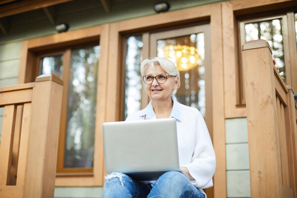 Smiling modern working senior lady in glasses sitting on porch of cottage and looking into distance while working on project using laptop