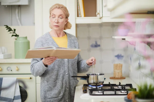 高级妻子一边看烹饪书里的食谱 一边站在煤气炉旁 在平底锅里煮东西 — 图库照片