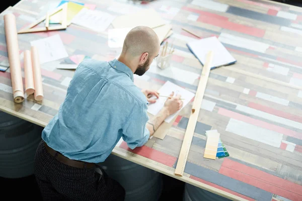 定規とカラースウォッチとスタジオで平らなプロジェクトを精緻化したロフトテーブルに傾いているひげを持つ集中ハゲ男性インテリアデザイナーの直接のビュー — ストック写真