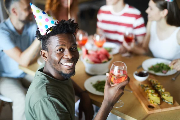 友人とお祝いのテーブルに座っている間 若いアフリカ系アメリカ人の男は カメラのために回転ガラスを飲みます — ストック写真