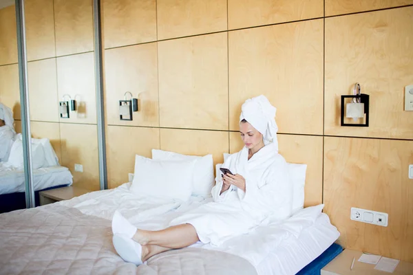 年轻的女性穿着白色浴衣 毛巾在头上 拖鞋坐在床上 并在智能手机上发短信 — 图库照片