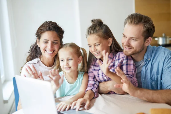 年轻而快乐的四口之家在笔记本电脑前围坐在桌旁 通过视频与亲朋好友聊天 — 图库照片