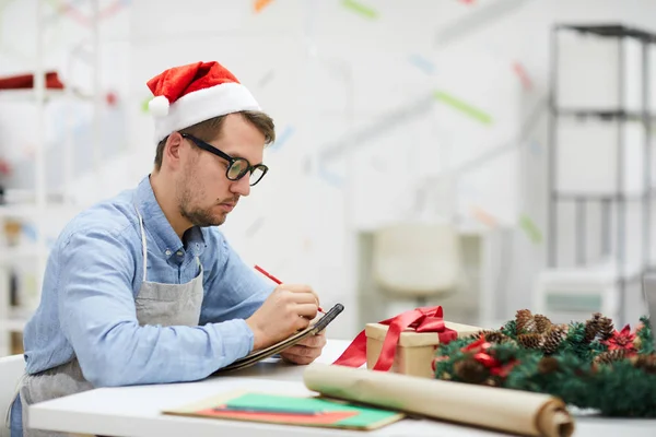 サンタの帽子とメガネで真面目な男性デザイナーがテーブルに座り スケッチパッドでメモを作りながら工芸スタジオで働きながら 彼は仕事を計画し 注文のリストを構成します — ストック写真