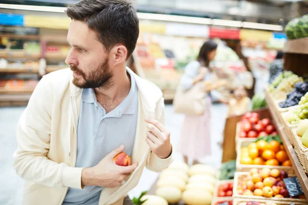 カジュアルな服装で真剣に心配したハンサムな若い男性泥棒が周りを見回し 食品店で果物を内側のポケットに入れて 彼はオーガニックショップで果物を盗みます — ストック写真