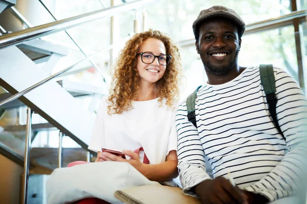 Feliz Joven Pareja Intercultural Mirándote Mientras Estás Sentado Escalera Dentro —  Fotos de Stock