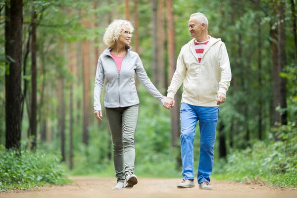 Senior Para Activewear Chodzenie Dół Forest Road Trzymając Ręce Patrząc — Zdjęcie stockowe