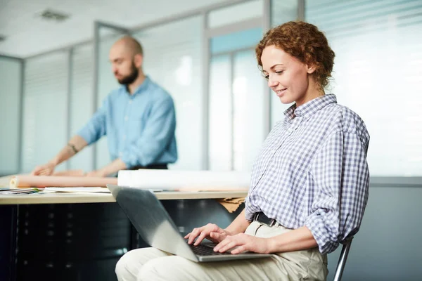 内容忙碌的女设计师在格子衬衫坐在椅子上 打字在笔记本电脑上 同时检查在线在办公室的 — 图库照片