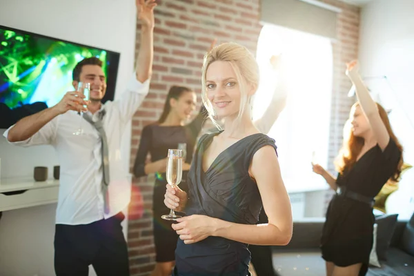 Posh blond female in black dress with flute of champagne looking at you while her friends dancing on background
