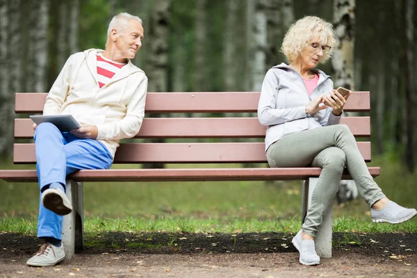 高级男子与触摸板看女性短信在智能手机在板凳的另一侧 — 图库照片