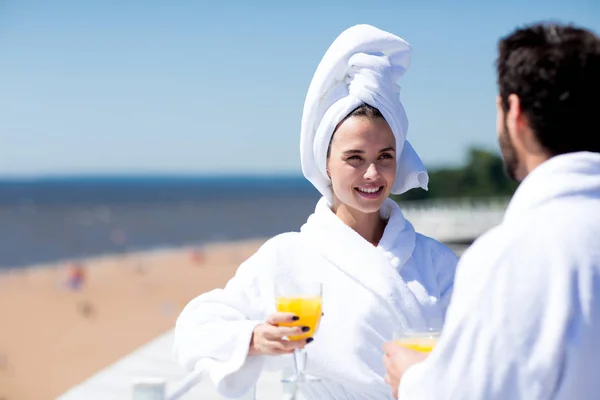 在避暑胜地度蜜月期间 穿着白色浴衣和毛巾的年轻女子喝果汁 并与丈夫交谈 — 图库照片