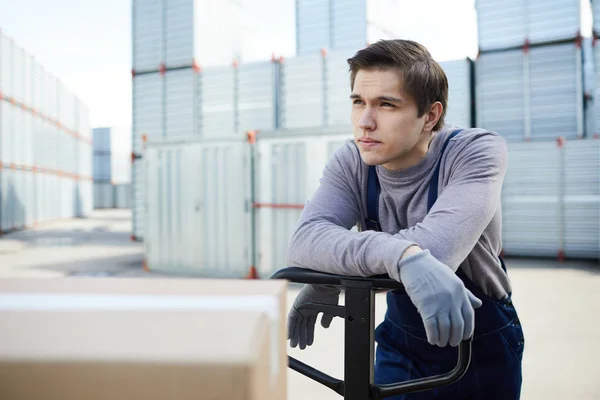 Young loader man leaning on handle of load cart and having minute of rest during work