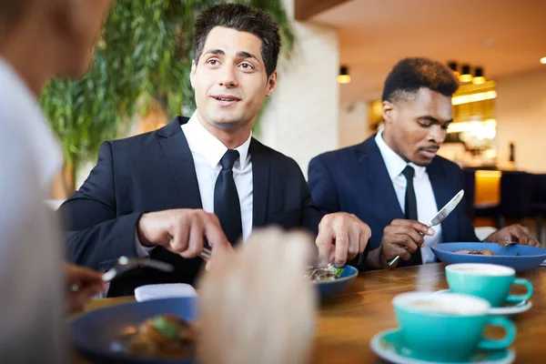 积极英俊的年轻商人穿着正式西装坐在桌旁 吃沙拉 同时在餐厅的晚餐与同事聊天 — 图库照片