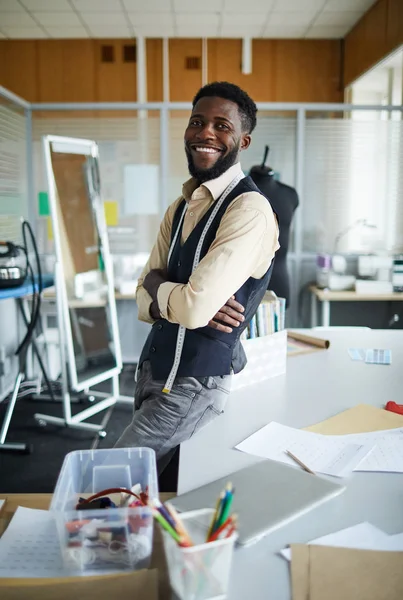 Cheerful African-american designer sitting on desk in his fashion studio or workshop