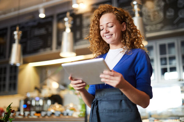 Young waitress with tablet reading online menu of restaurant or cafe during work