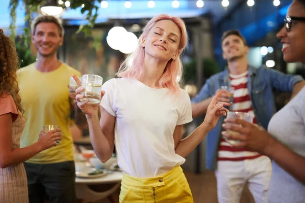 カジュアルウェアで興奮した女の子は 彼女の友人の背景に飲み物やダンスを保持 — ストック写真