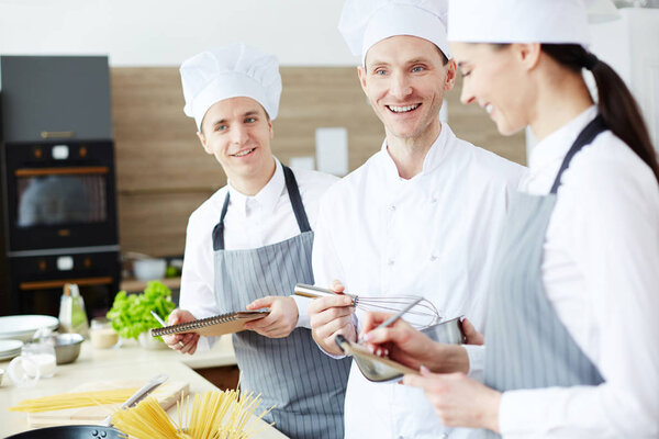 Positive excited handsome chef whipping ingredients with whisk while enjoying cooking with young intern employees at commercial kitchen