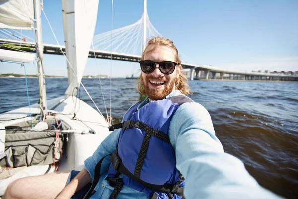 快乐兴奋的英俊的年轻人 留着胡子 戴着太阳镜笑着 一边拍摄自己对城市桥 而漂浮在游艇上 — 图库照片
