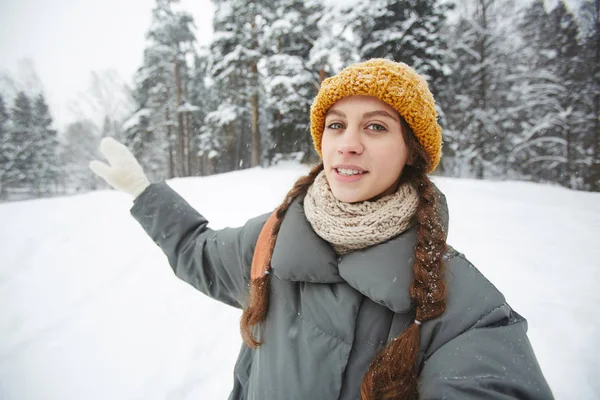 微笑内容年轻的女性旅游博客在针织帽子和围巾手势的手 同时显示森林和在冬季森林旅游 她看着相机 — 图库照片