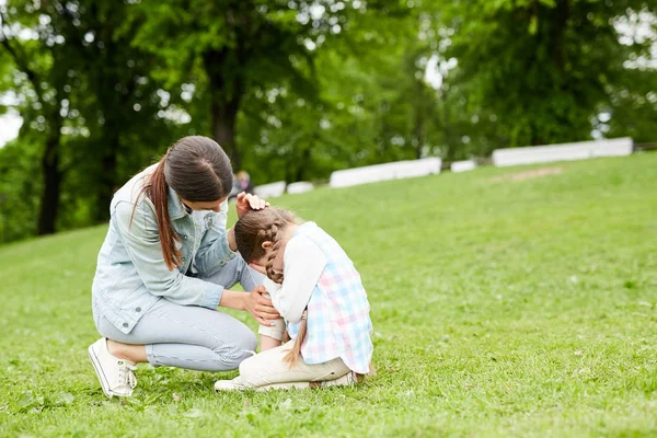 緑の芝生の上に座っている間に泣いている小さな女の子と彼女を慰める彼女の母親 — ストック写真