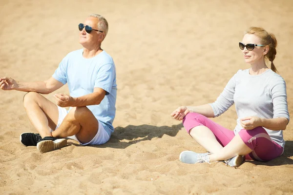 现代高级夫妇穿着运动服和太阳镜 在锻炼后坐在沙滩上摆出莲花的姿势 — 图库照片