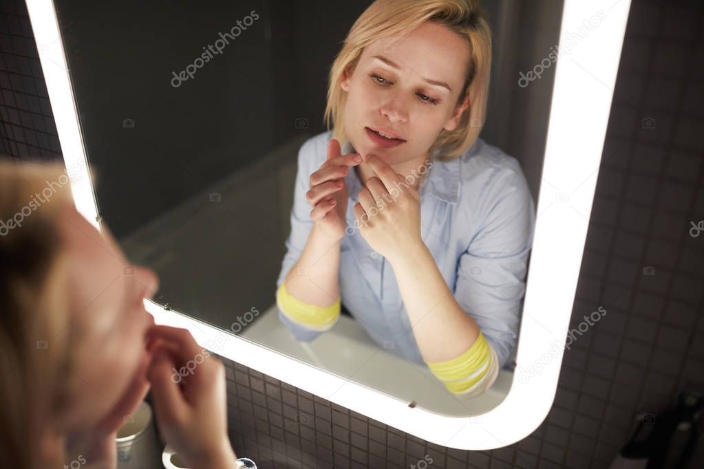 Young attractive blond Caucasian woman popping pimple on her chin while looking at herself in bathroom mirror