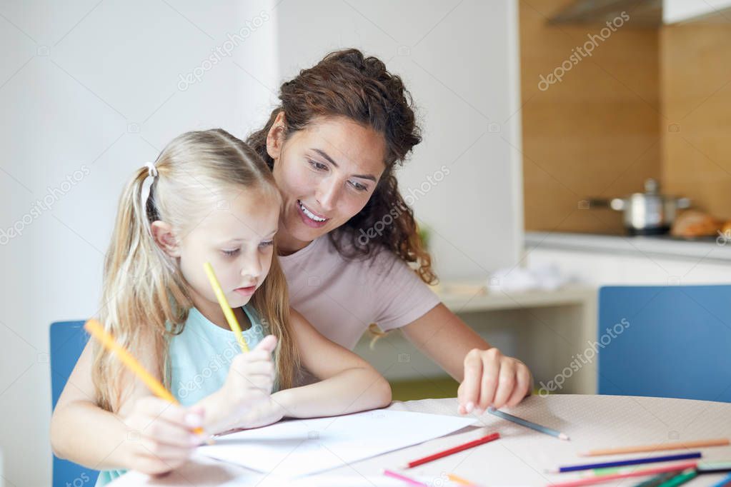 Young smiling woman and her little daughter with crayons sitting by table and thinking of what to draw