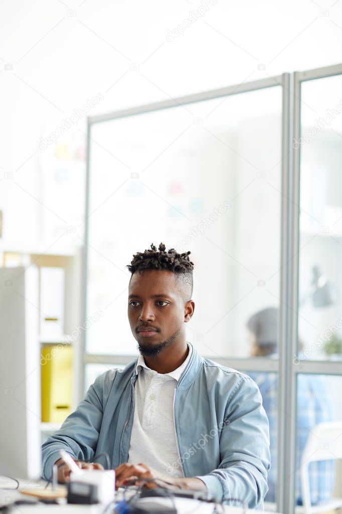 Young African-american hipster sitting by workplace in front of computer monitor and learning new code language
